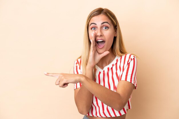 Chica rubia uruguaya aislada sobre fondo beige apuntando hacia el lado para presentar un producto y susurrando algo