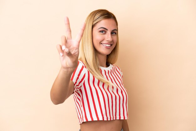 Chica rubia uruguaya aislada sobre fondo beige sonriendo y mostrando el signo de la victoria