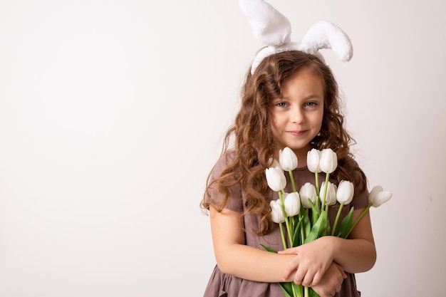 una chica rubia con un vestido de algodón marrón y orejas de conejo de Pascua sostiene un ramo de tulipanes blancos