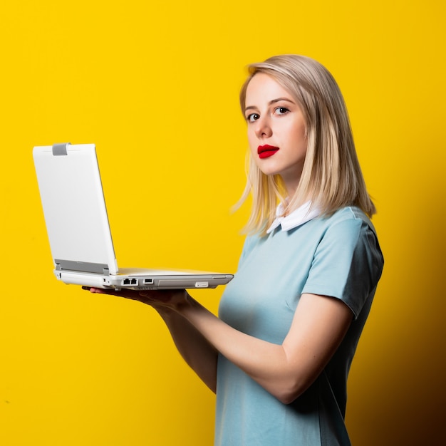 Chica rubia en vestido azul con computadora portátil en espacio amarillo