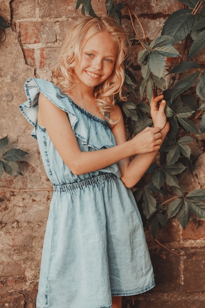 Una chica rubia con un vestido azul está de pie contra la pared con uvas silvestres. Foto de alta calidad