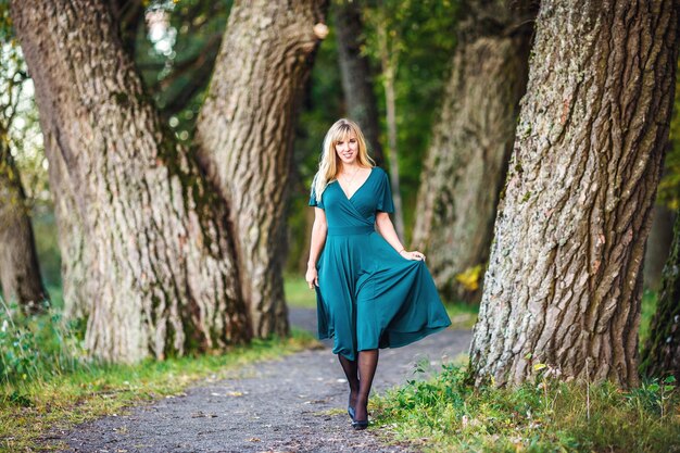 Una chica rubia con un vestido largo verde camina por un robledal