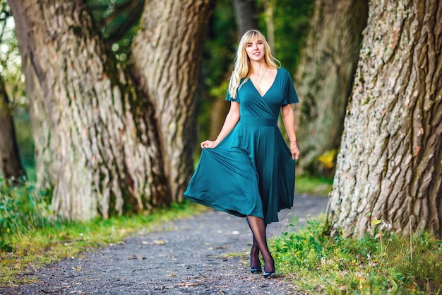 Una chica rubia con un vestido largo verde camina por un robledal