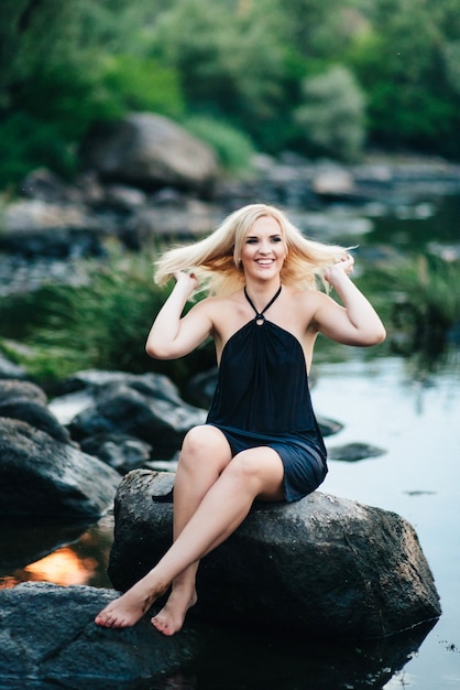 Chica rubia con un vestido negro con ojos azules en la costa de piedra del río