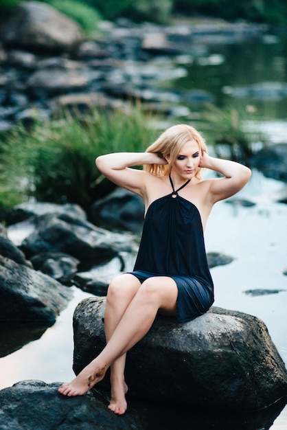 Chica rubia con un vestido negro con ojos azules en la costa de piedra del río