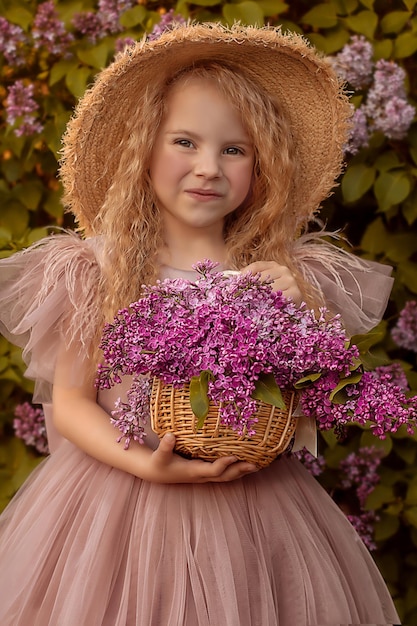 Una chica rubia con un vestido de pelo largo con un sombrero de paja sostiene una canasta de lilas en sus manos