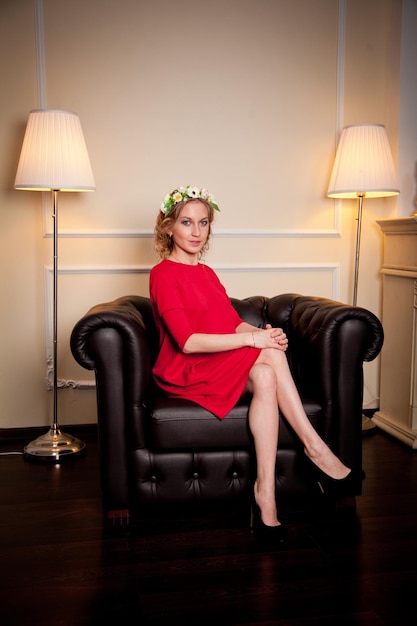 Una chica rubia con un vestido rojo y flores en el pelo se sienta en un sofá de cuero