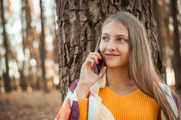 Chica rubia vistiendo un suéter naranja cubierto de cuadros brillantes hablando por teléfono en el parque