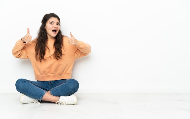 Chica rusa adolescente sentada en el suelo dando un pulgar hacia arriba gesto