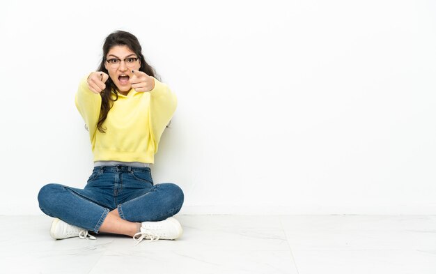 Chica rusa adolescente sentada en el suelo sorprendida y apuntando hacia el frente