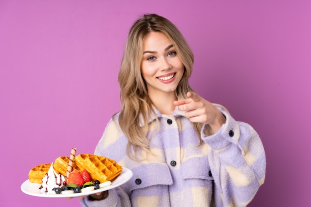 Chica rusa adolescente sosteniendo gofres en la pared púrpura apuntando hacia el frente y sonriendo