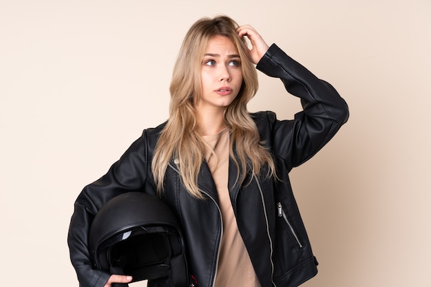 Chica rusa con un casco de moto en la pared de color beige que tiene dudas mientras se rasca la cabeza