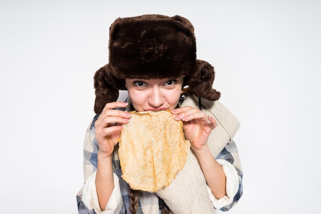 Chica rusa divertida en un sombrero de piel negro come panqueques calientes celebra Shrovetide