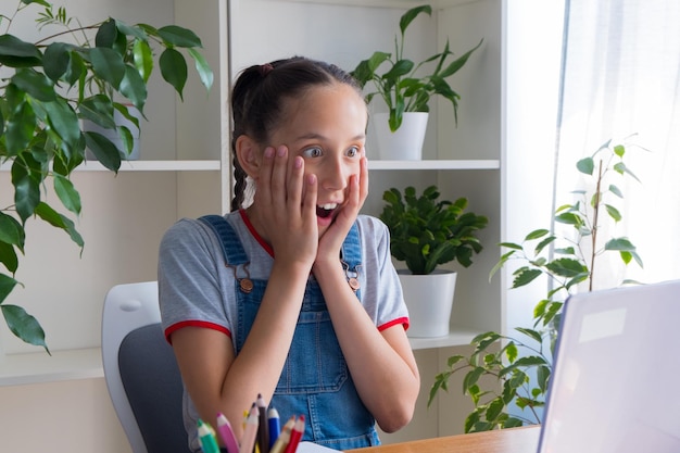 Una chica sentada en casa estudiando hablando por Internet con alegría y con admiración mira la pantalla del portátil