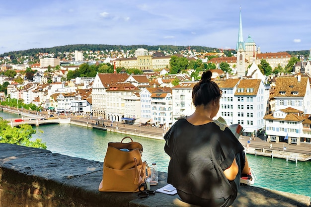 Chica sentada en la colina de Lindenhof y mirando una guía en Zúrich, Suiza. Limmatquai y Predigerkirche al fondo