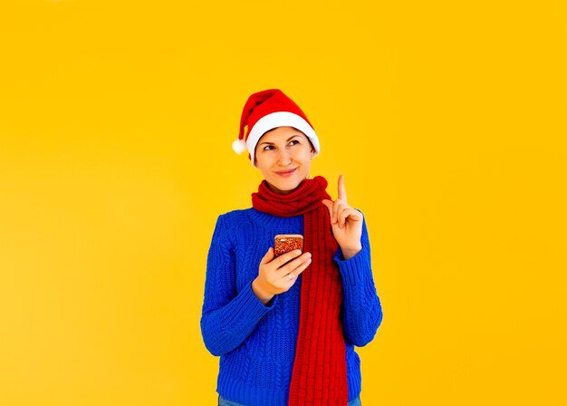 Chica sentada en Internet en Navidad usando un teléfono inteligente. Una niña con un suéter azul y gorro de Papá Noel mira el teléfono sobre un fondo amarillo. tiro del estudio