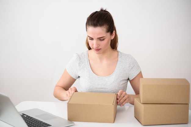 Chica sentada en una mesa de ordenador blanca con un ordenador portátil con interés abre una caja de cartón