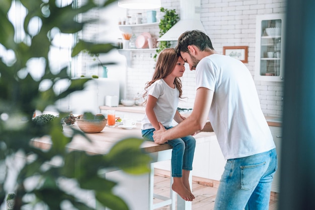 Chica sentada en la mesa con el padre parado cerca de la vista de la cocina detrás de la planta en maceta