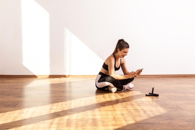 Chica sentada en el suelo con una tableta buscando video tutorial para hacer ejercicio deportivo en casa con top deportivo negro y medias Foto de estudio de longitud completa iluminada por la luz del sol desde la ventana