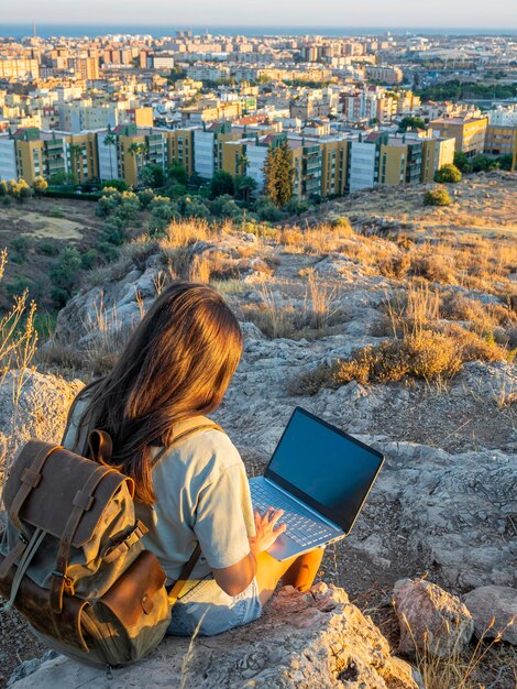 Chica sentada teletrabajando con un portátil nómada digital viajando Al fondo la ciudad de Málaga