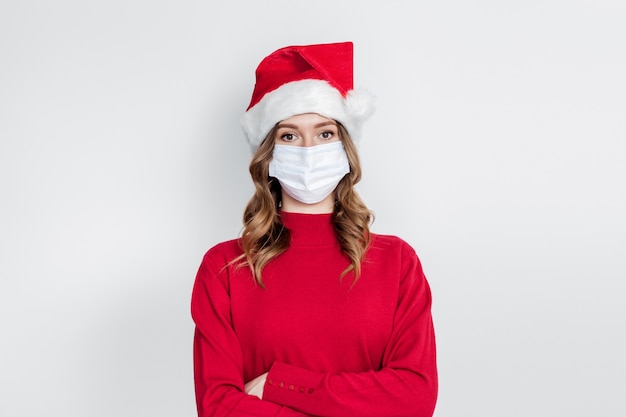 Una chica seria con una gorra de Año Nuevo, un suéter rojo con una máscara médica se encuentra aislado en un fondo blanco. Concepto de celebración de año nuevo 2021