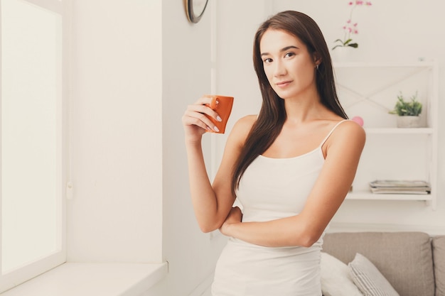 Chica seria parada cerca de la ventana en casa, tomando café, copiando espacio