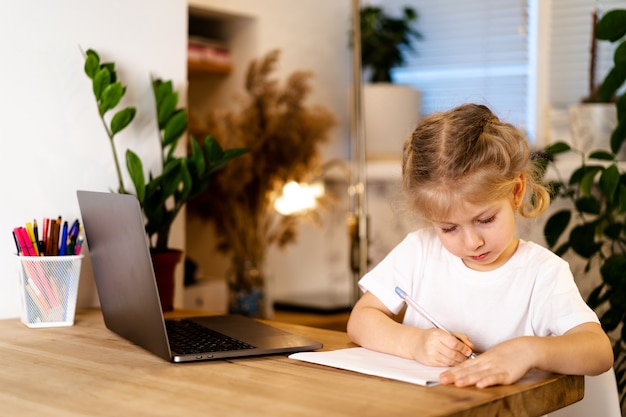 Foto chica seria riéndose de su computadora portátil y haciendo los deberes en un cuaderno