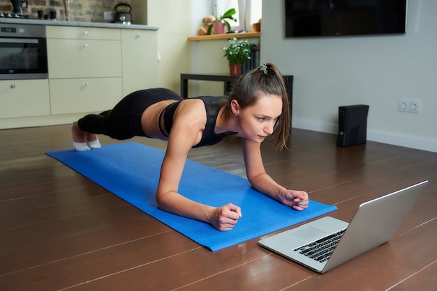 Una chica seria con un traje ajustado de entrenamiento negro está haciendo plancha viendo un video en línea en una computadora portátil