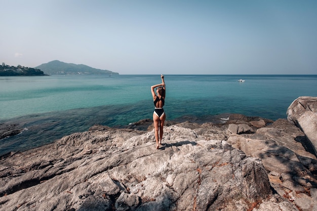 Chica sexy en bikini negro de pie en la cima de la montaña sobre el mar azul y vistas al barco. Phuket. Tailandia Hermoso paisaje marino. Vista trasera