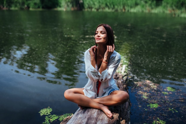 Una chica sexy con una camisa blanca se sienta en una madera flotante en posición de loto cerca del río. El concepto de recreación al aire libre.