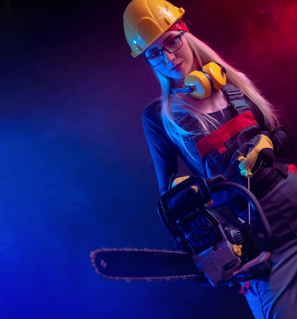 Foto chica sexy con una motosierra en un mono de construcción y casco sobre un fondo negro con luz de neón