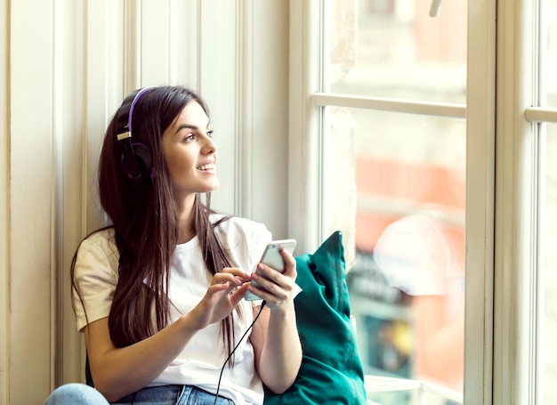 Chica sexy de pelo largo usa auriculares y camiseta blanca escucha música sentada en el alféizar de la ventana