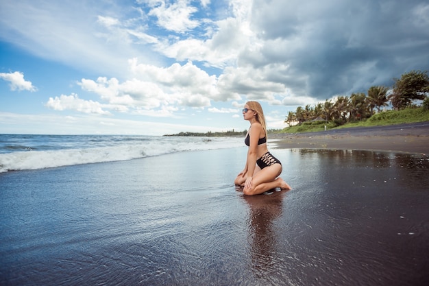 Chica sexy en traje de baño se sienta en la playa con arena negra