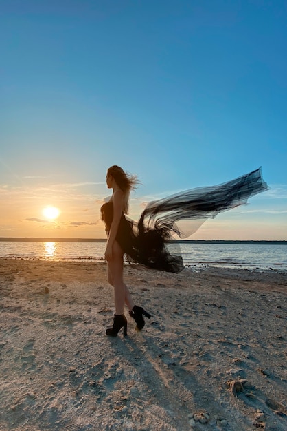 Chica sexy con un vestido negro en la playa al atardecer.