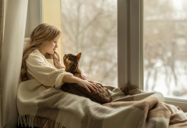 La chica se sienta en el alféizar junto a la ventana con un gato.