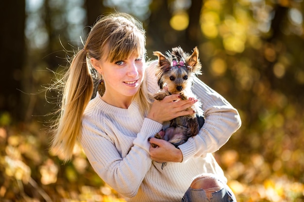 La chica se sienta y se relaja en el suelo en el bosque de otoño y juega con el perro pequeño Yorkshire Terrier