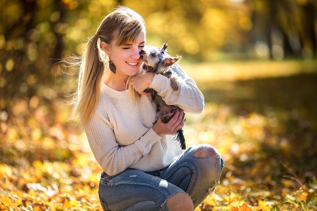 La chica se sienta en el suelo en el bosque de otoño y juega con un perro pequeño de Yorkshire Terrier