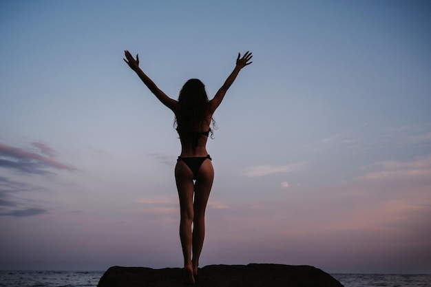 Chica de silueta con un botín sexy de pie sobre las rocas en la playa junto al mar