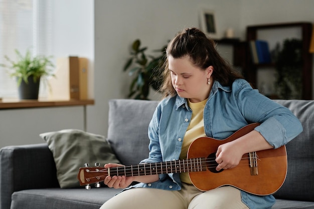 Chica con síndrome de down aprendiendo a tocar la guitarra mientras se sienta en el sofá en la sala de estar