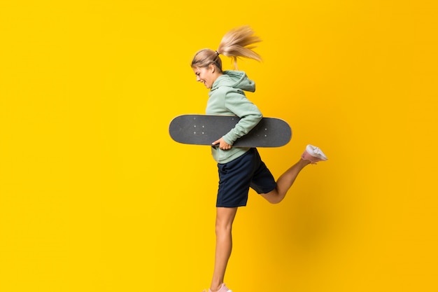 Chica skater adolescente rubia saltando sobre fondo amarillo aislado