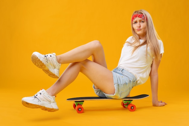 Chica skater en pantalones cortos y camiseta sentado a bordo sobre fondo amarillo