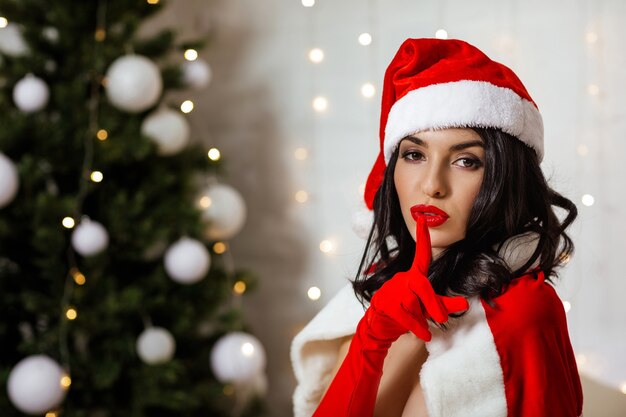 Chica Snata con el dedo en sus labios rojos mostrando Shh en el fondo del árbol de Navidad