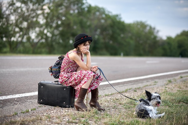 Chica solitaria con perro y maleta de pie sobre la carretera