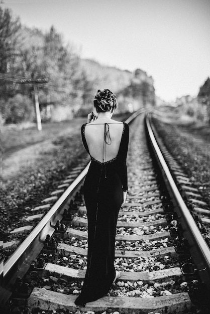 Chica solitaria con un vestido negro camina con la espalda en las vías del tren