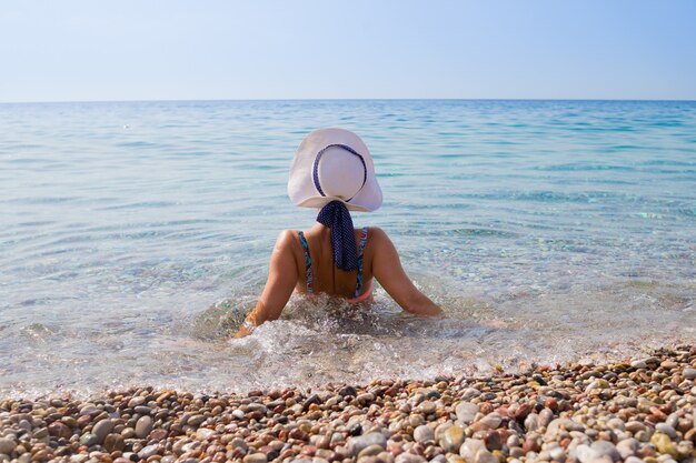 Chica con sombrero blanco sentado a la cámara sobre guijarros en la playa