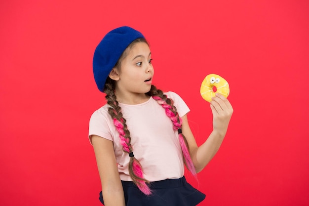 Chica con sombrero de boina sostenga donut fondo rojo Niño niña juguetona come donut Concepto de salud y nutrición Vida dulce Concepto de tienda de dulces y panadería Niño fanático de donuts horneados Donut dulce delicioso