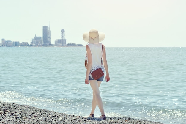 Chica con sombrero camina por la orilla del mar. Ciudad a lo lejos. Vista desde atrás