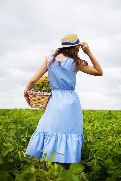 Chica con sombrero camina sobre un campo verde con una canasta de flores.