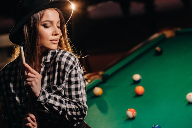 Una chica con un sombrero en un club de billar con un taco en sus manos. Juego de billar.