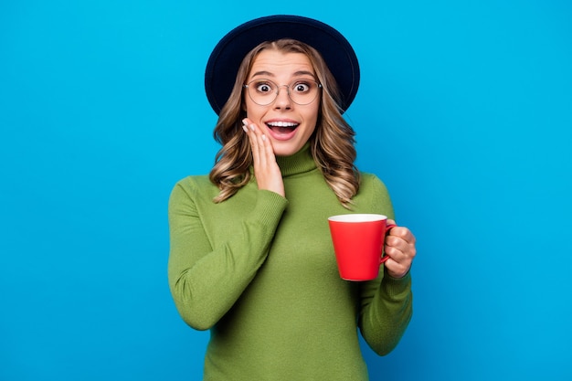 Chica con sombrero y gafas sosteniendo taza aislado en azul